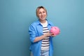 blond attractive young woman in casual attire thoughtfully holding a piggy bank with money on a blue background with Royalty Free Stock Photo