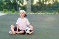 A blond boy in a cap in a sports uniform sits on a football field with a soccer ball, sports section. Training of children, Royalty Free Stock Photo