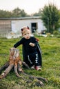 A blond boy in a devil costume with a red cloak stands near a tree stump. Halloween eve, carnival costume, traditions Royalty Free Stock Photo