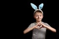 Blond boy in hare ears folded his hands in heart shape on his chest on black background. Love and family concept. Copy space