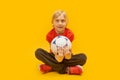 Blond boy hugs the ball with sits cross-legged in isolation on yellow background. Child footballer