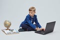 Blond boy in a school suit is sitting in front of a laptop, a globe, and a book. photo session in the Studio on a white background