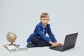 Blond boy in a school suit is sitting in front of a laptop, a globe, and a book. photo session in the Studio on a white background