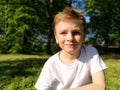 Blond boy 7 years old in a white T-shirt on the background of a forest. The child looks to the side, squints his eyes from the