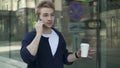 Young man with coffee walking along building and talking on phone