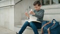 Blond child student writing on book breaking paper sitting on stairs at school