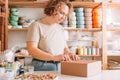 Blond confident woman packing and wrapping parcel with silicone baby dishes in cardboard box package, stamping cardboard