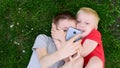 Blond and dark-haired boys lay on grass, goofing around and taking selfies. Top view.