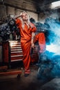 Blond female mechanic with tattooed hands in orange overalls stands in garage or workshop