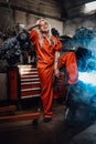 Blond female mechanic with tattooed hands in orange overalls stands in garage or workshop