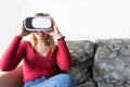 Blond girl with futuristic experience, enjoying sitting on the coach at home, playing a video game with virtual reality glasses