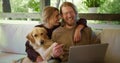A blond girl and a blond guy with a beard in glasses are looking at something funny on a laptop and laughing next to