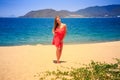 blond girl in red stands barefoot on sand beach at noon