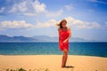 blond girl in red stands on sand holds hand on hip at noon