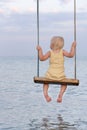 Blond girl sitting on rope swing over the water. Back view. Happy childhood Royalty Free Stock Photo