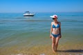 A blond girl wearing sunglasses and a white hat is standing on the beach opposite the yacht. Royalty Free Stock Photo