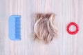 Blond hair lock, red scrunchie, blue plastic comb light wooden background close up, cut off blonde hair curl on bright wood, brush Royalty Free Stock Photo
