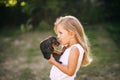 Blond kid girl in white dress is kissing puppy pet dog outdoors. Side view Royalty Free Stock Photo