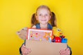 Blond little girl holding a box with toys on yellow background. Donation concept.. Royalty Free Stock Photo