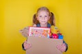 Blond little girl holding a box with toys on yellow background. Donation concept.. Royalty Free Stock Photo