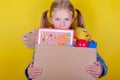 Blond little girl holding a box with toys on yellow background. Donation concept.. Royalty Free Stock Photo