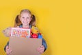 Blond little girl holding a box with toys on yellow background. Donation concept.. Royalty Free Stock Photo