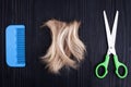 Blond lock of hair, metal scissors, plastic comb black wooden background closeup, cut off blonde hair curl on dark wood, shears