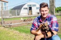 Blond man holding a baby goat in front of a barn