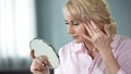 Blond middle-aged lady looking in mirror at her eye wrinkles, beauty injections