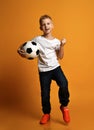 Blond positive boy child in white t-shirt and jeans standing with soccer ball in hand and showing win sign with hand