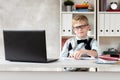 Blond schoolboy wearing sweater and glasses sits behind desk and watching at laptop