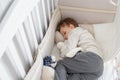 Blond seven years boy sleeping in his sister white wooden cot in white shirt and grey trousers with toys in his arms