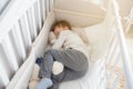 Blond seven years boy sleeping in his sister white wooden cot in white shirt and grey trousers with toys in his arms