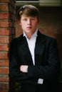 A blond teen in a tuxedo leaning on a brick wall.
