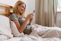 Blond woman smiling and using cellphone while lying in bed at home Royalty Free Stock Photo