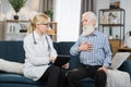 Blond woman doctor in glasses and white medical coat, visits her senior patient at home. Therapist is taking notes on Royalty Free Stock Photo
