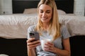 Blond woman drinking tea and using cellphone sitting over bed at home Royalty Free Stock Photo