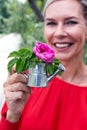 Blond woman outdoors holding a miniature watering can with a flower