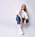 Blonde adolescent in sunglasses, watch, jeans, sweater, socks and sneakers. She squatting on floor, isolated on white. Close up Royalty Free Stock Photo