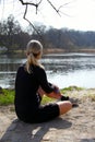 Blonde athlete girl sitting on ground to relax after jogging with water bottle under a tree on a lake shore lit by sunlight - Runn