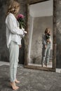 Blonde beautiful model with curly hair look in the mirror. Girl holding pink flower in studio