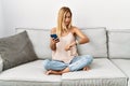 Blonde beautiful young woman sitting on the sofa at home using smartphone checking the time on wrist watch, relaxed and confident Royalty Free Stock Photo