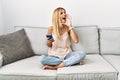 Blonde beautiful young woman sitting on the sofa at home using smartphone shouting and screaming loud to side with hand on mouth Royalty Free Stock Photo