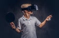 Blonde boy dressed in a white t-shirt playing ping-pong with a virtual reality glasses. Isolated on dark textured