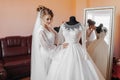 A blonde bride in a long lace robe and white shoes stands in her room next to her wedding dress, posing by the mirror. Royalty Free Stock Photo