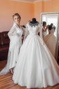 A blonde bride in a long lace robe and white shoes stands in her room next to her wedding dress, posing by the mirror. Royalty Free Stock Photo