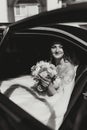 A blonde bride, smiling sincerely, sits in a black car on her wedding day with a bouquet of white roses. Royalty Free Stock Photo