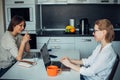 Blonde and brunette with laptops in kitchen area. Two cute woman working, sitting at table facing each other in a relaxed home Royalty Free Stock Photo