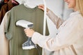 Blonde caucasian female using electric steamer at home, ironing clothes on hanger