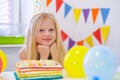 Blonde caucasian girl sits thoughtfully and dreamily at festive table near birthday rainbow cake and makes a wish Royalty Free Stock Photo